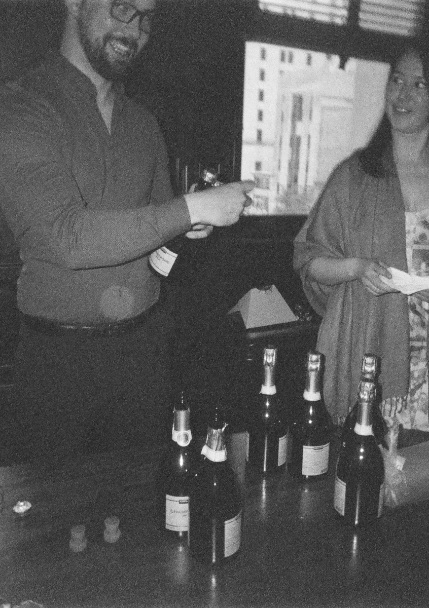 A man in a gray shirt prepares to open a champagne bottle, while a woman smiles nearby, surrounded by several bottles of champagne on a table.