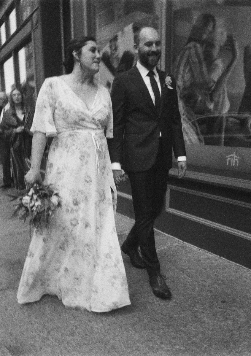 A bride in a floral dress and a groom in a suit walk hand in hand on a city street, surrounded by guests in the background.