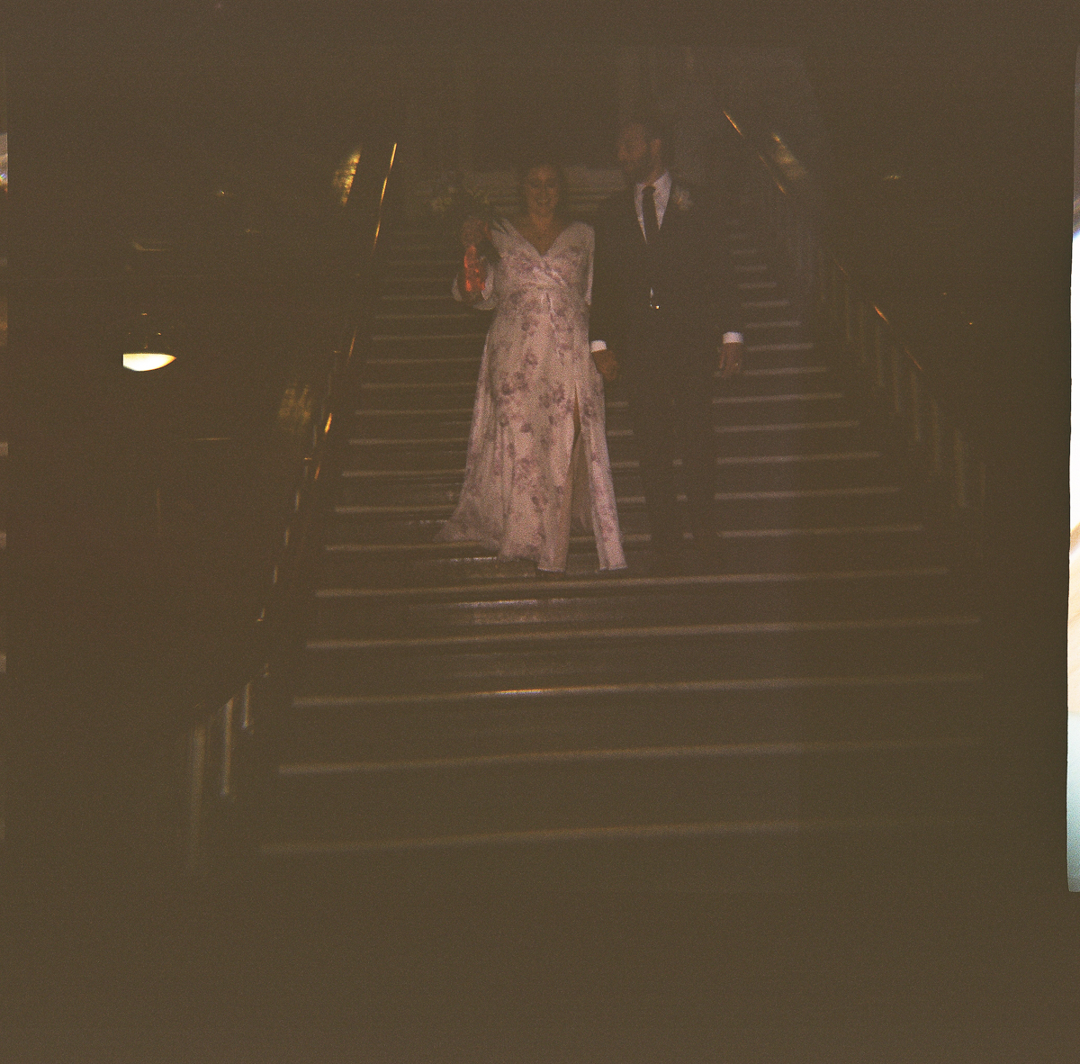 A bride in a floral gown and a groom in a suit holding hands while walking down a set of illuminated stairs.