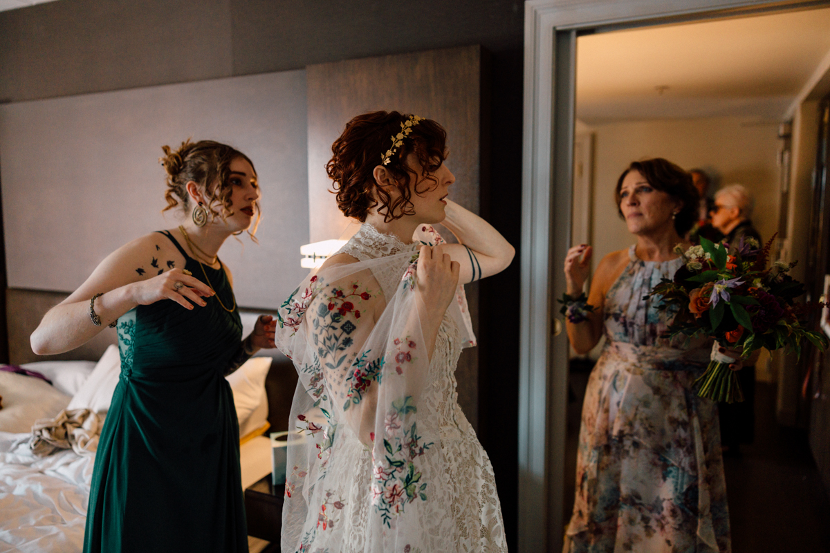 A bride adjusts her veil while two women stand nearby, one helping with her dress and the other holding a bouquet, in a stylish hotel room setting.
