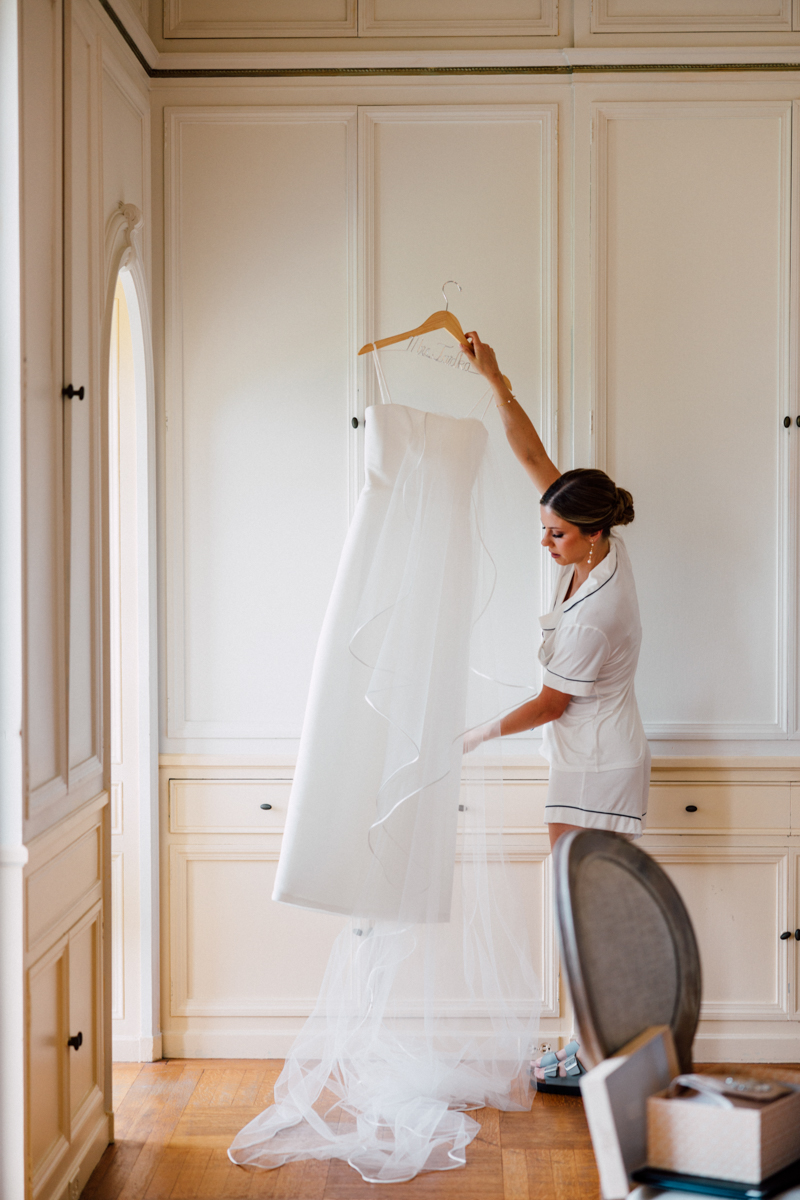 A woman in a white outfit gracefully holds a wedding dress with a veil in a beautifully lit room, surrounded by classic interior decor.