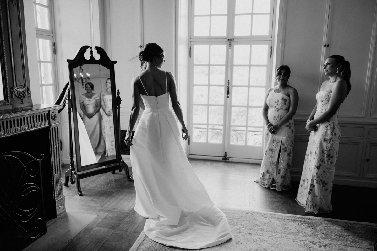 A bride in a white gown looks at her reflection in a mirror, while two women in floral dresses observe her in a beautifully lit room.