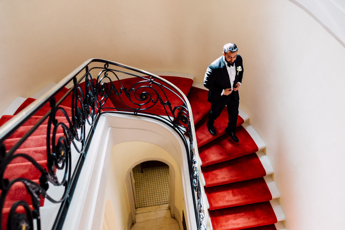 A groom in formal attire descends a spiral staircase with a lush red carpet, adorned with stylish black railings, exuding sophistication.