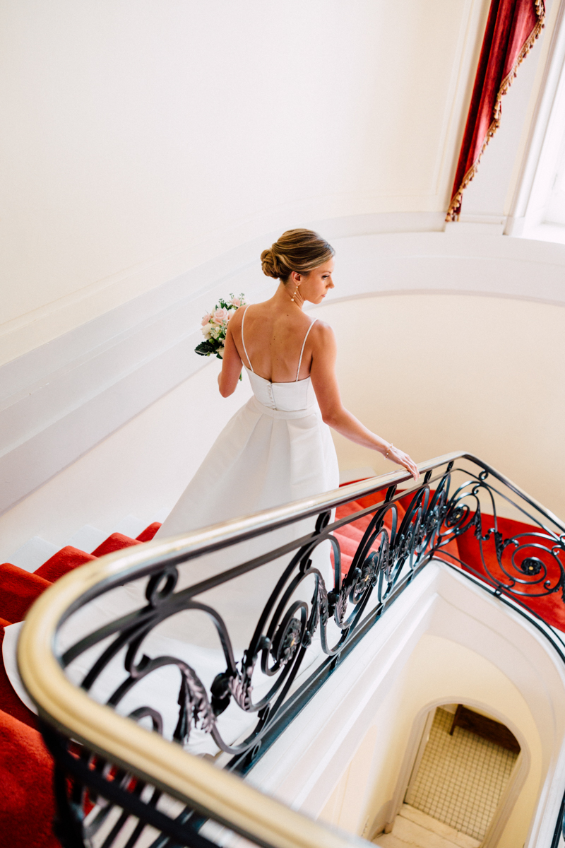 A bride in a simple white dress climbs a stylish staircase while holding a bouquet, showcasing a serene and graceful moment.