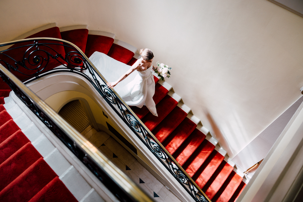 A bride in a white gown gracefully climbs a lavish staircase adorned with rich red carpet, holding a bouquet of flowers.