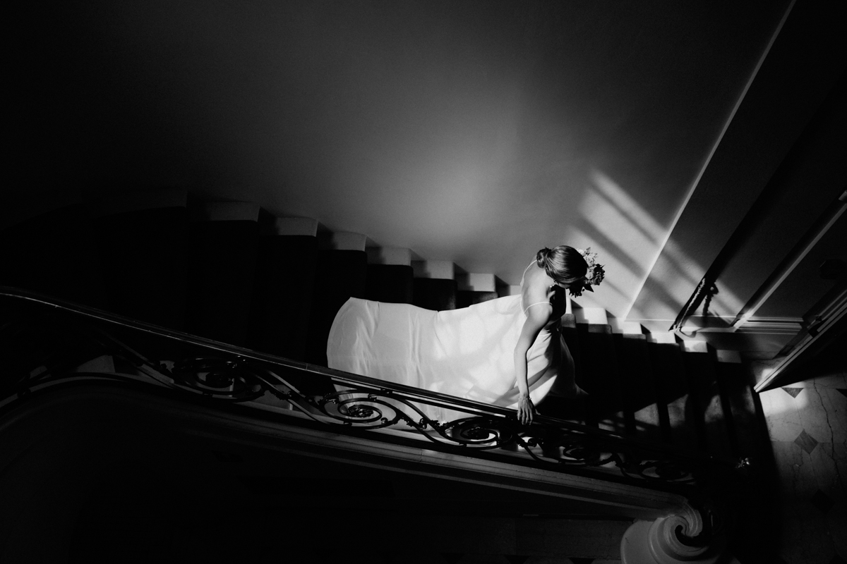A bride gracefully descends a staircase in a flowing white gown, illuminated by soft light and shadows.