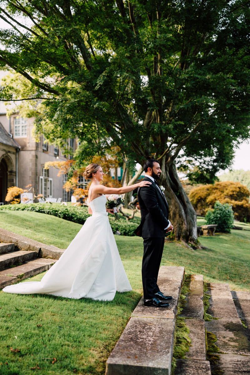 A bride approaches the groom from behind, preparing for their emotional first look amidst a beautiful outdoor setting with greenery and elegant architecture.