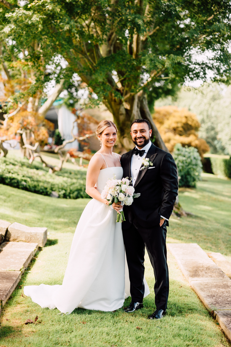 A joyful couple poses together in a beautifully landscaped garden, the bride wearing a classic white gown and holding a bouquet, while the groom is dressed in a formal black tuxedo.