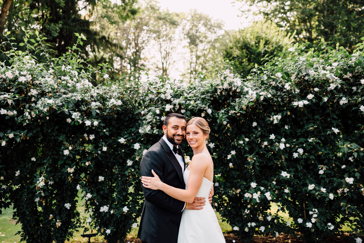 A joyful couple stands closely together, smiling in front of a lush backdrop of white flowers and greenery.