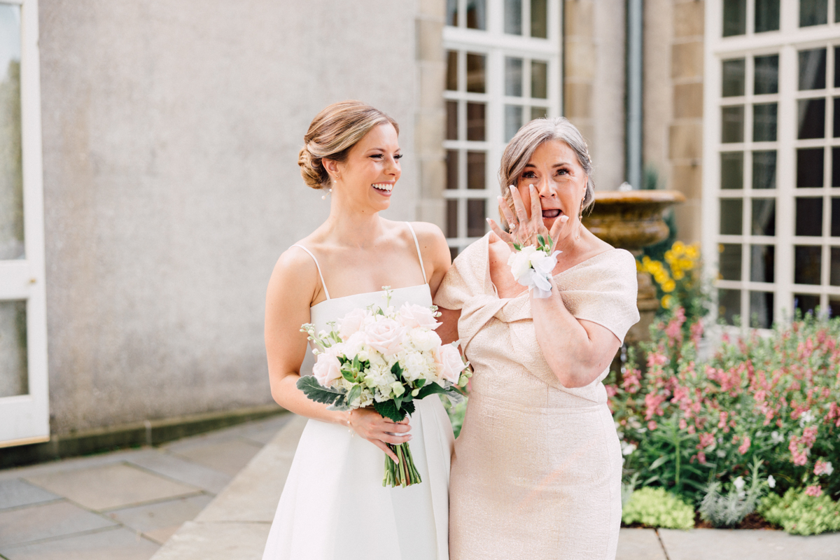 A happy bride and her mother share an emotional moment, surrounded by a lush garden, as the bride holds a bouquet of flowers.