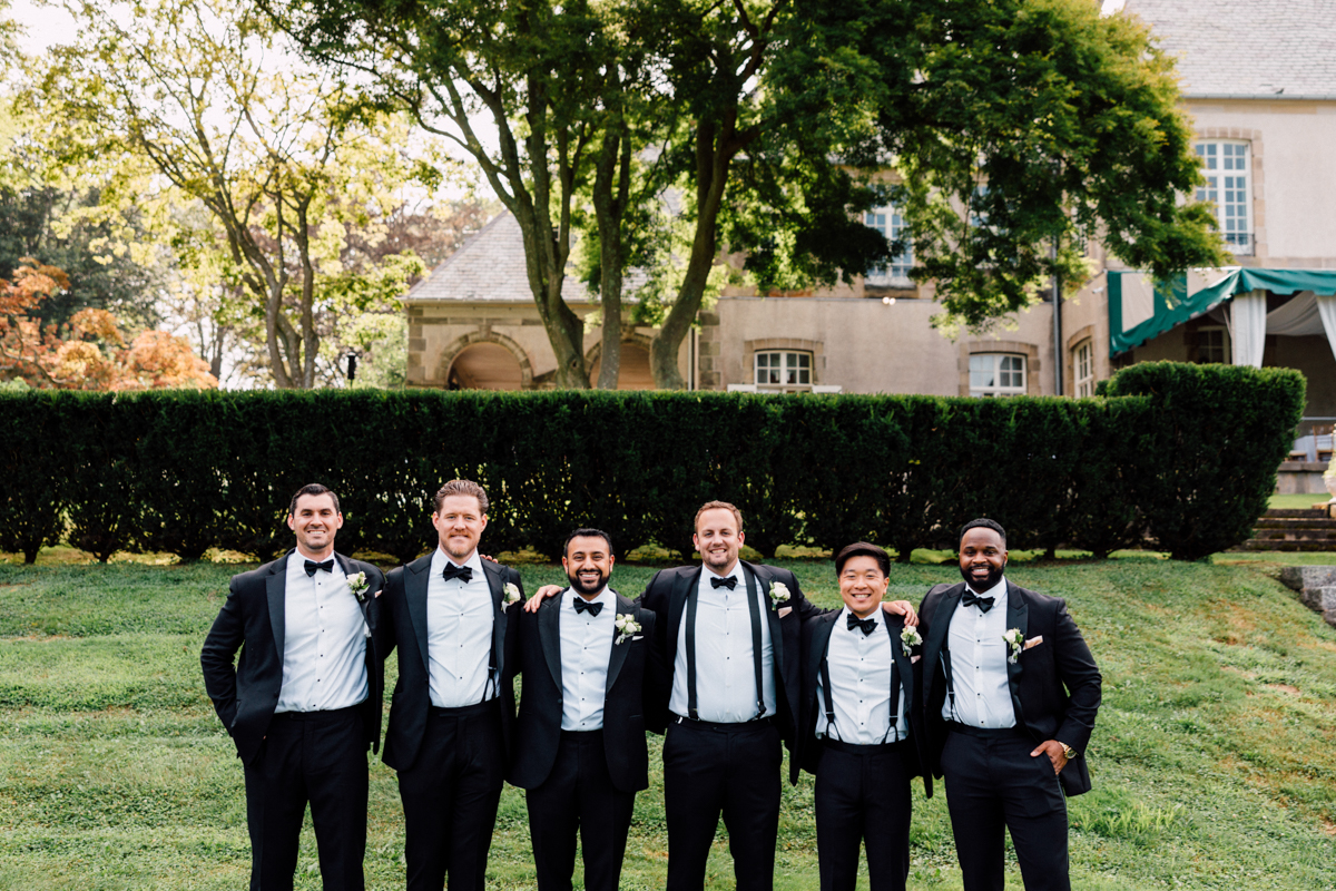 A group of six men in formal black tuxedos and bow ties standing together outdoors in front of a manicured hedge and a historic building.