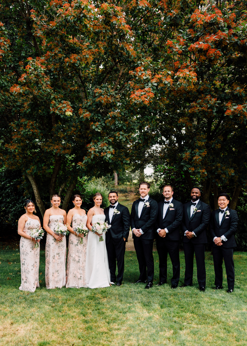 A wedding party posed elegantly outdoors among colorful foliage, featuring bridesmaids in floral dresses and groomsmen in tuxedos.