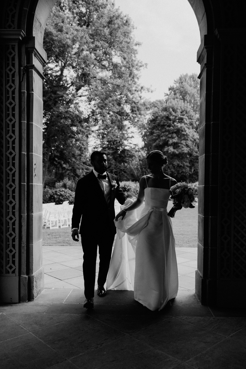 A bride and groom stepping out of an archway, framed by lush greenery and soft light, capturing a moment of joy and anticipation.