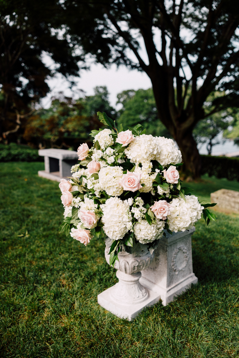 A beautifully arranged bouquet of white hydrangeas and pink roses in a stone planter, set against a lush green garden backdrop.
