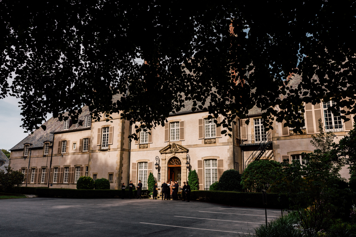 A large, stylish mansion surrounded by lush greenery, with guests gathered at the entrance and a clear sky above.
