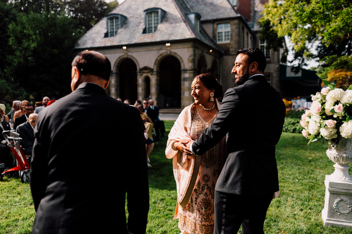 A joyful woman in traditional attire shares a smile with a man as they walk through a garden setting during a wedding ceremony, surrounded by guests.