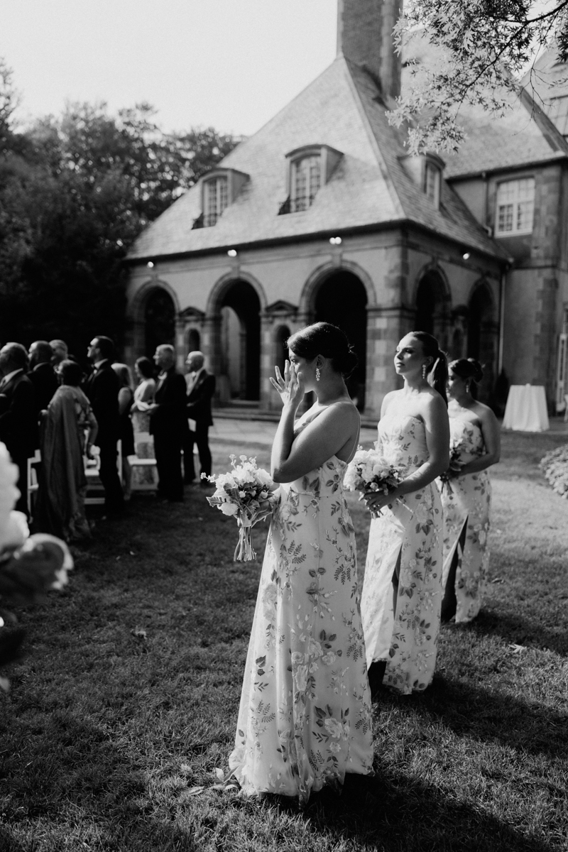 Bridesmaids wearing floral dresses stand in a garden during a wedding ceremony, with one touching her face and others holding bouquets.