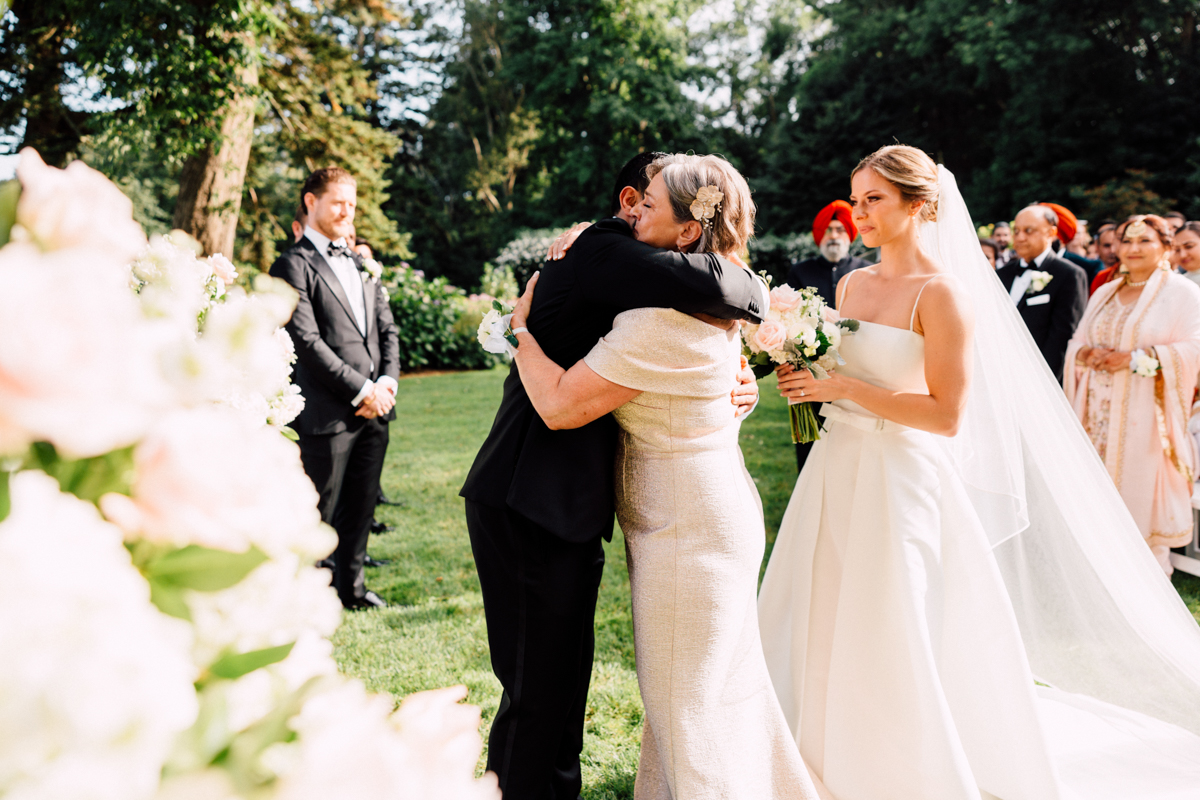 A bride watches as her partner embraces a family member, surrounded by a lush garden and guests.