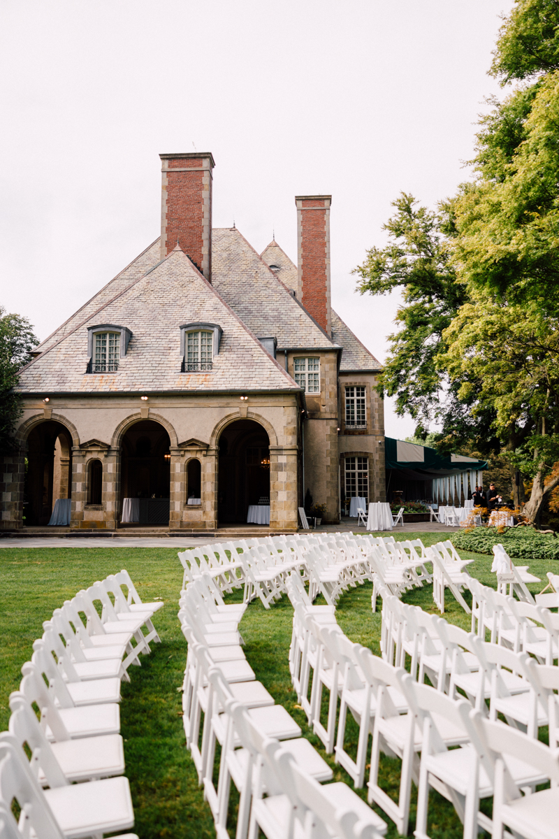 A picturesque mansion with chimneys and distinctive architecture, surrounded by rows of white chairs set up on a well-manicured lawn for a wedding ceremony.