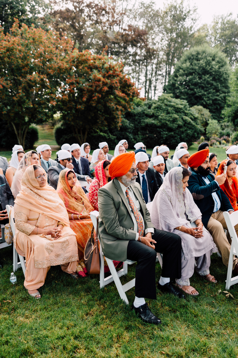 A gathering of Sikh individuals seated outdoors, dressed in traditional attire, as they participate in a religious or cultural ceremony surrounded by lush greenery.