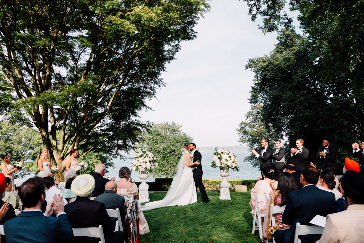 A couple kisses during their outdoor wedding ceremony by the lake, surrounded by guests and floral arrangements.