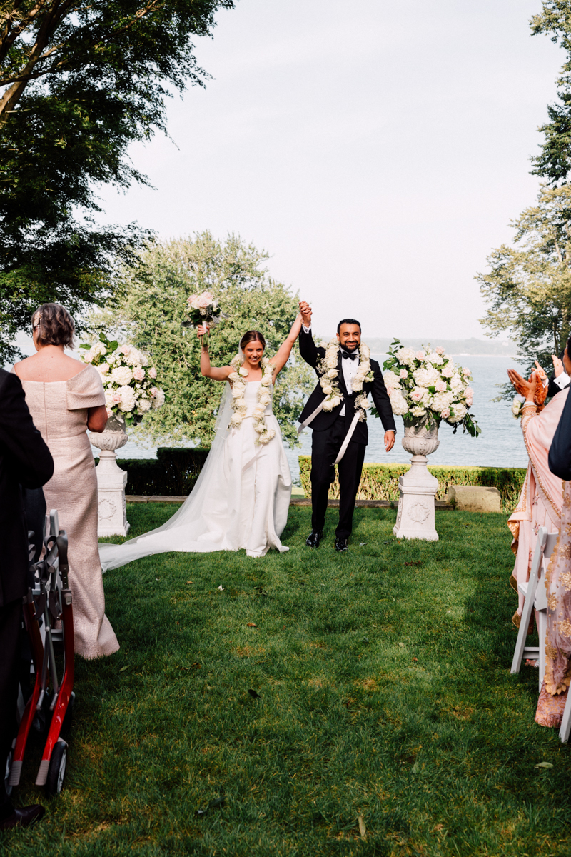 A bride and groom joyfully raise their hands together after their wedding ceremony, surrounded by floral arrangements and a scenic backdrop.
