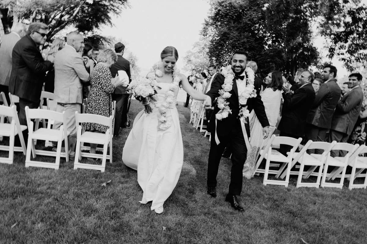 A smiling bride and groom walk hand in hand down the aisle, adorned with floral garlands, as guests celebrate their union.