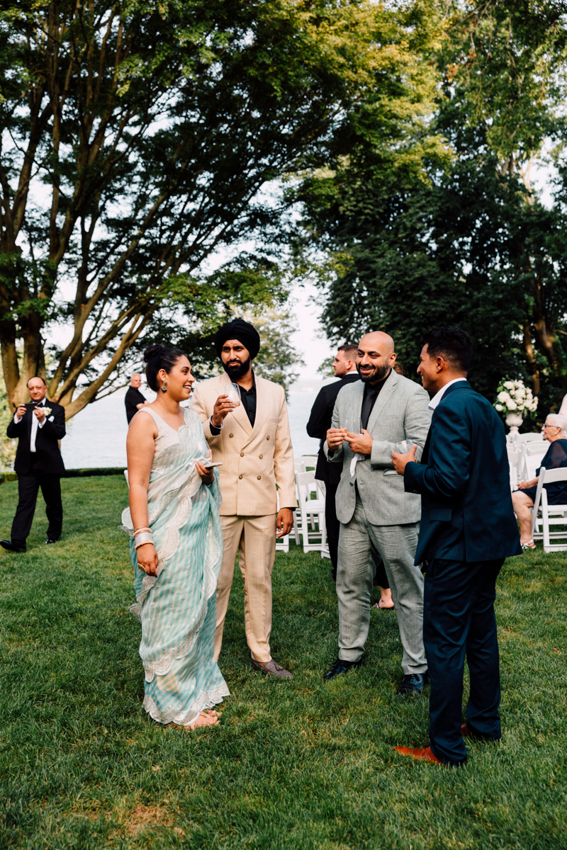 A group of four individuals, dressed in elegant attire, gather outdoors, enjoying drinks and laughter in a scenic park setting with trees and a view of the water in the background.