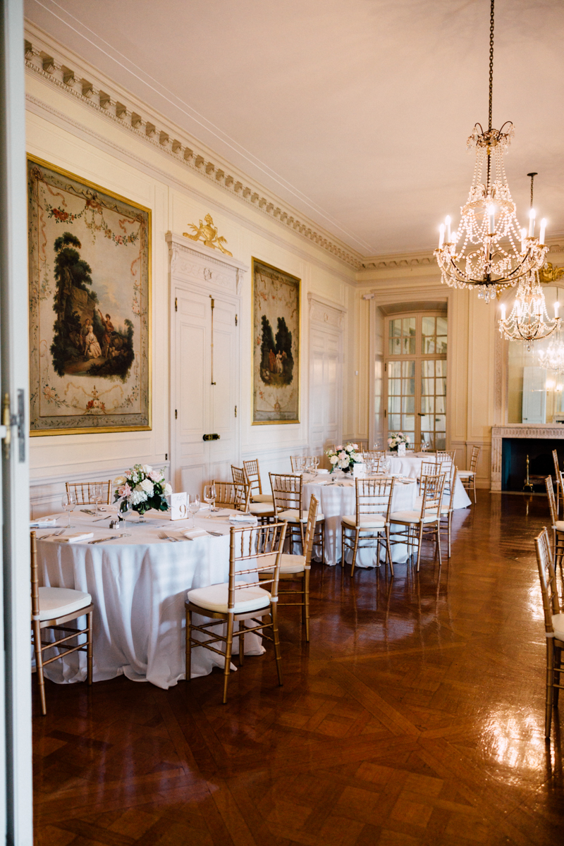 A beautifully arranged dining room featuring elegant tables with white linens, gold-framed chairs, and floral centerpieces, set in a sophisticated space adorned with classic paintings and chandeliers.