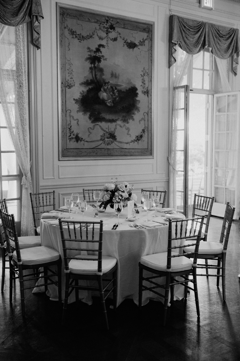 A round table elegantly set with glassware, silverware, and floral arrangements in a vintage-style dining room featuring large windows and classic artwork on the walls.