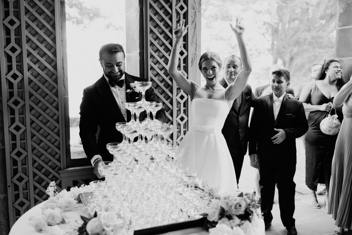 A joyful bride raises her arms in celebration beside a man pouring champagne into a tower of glasses, with guests enjoying the festivities in the background.