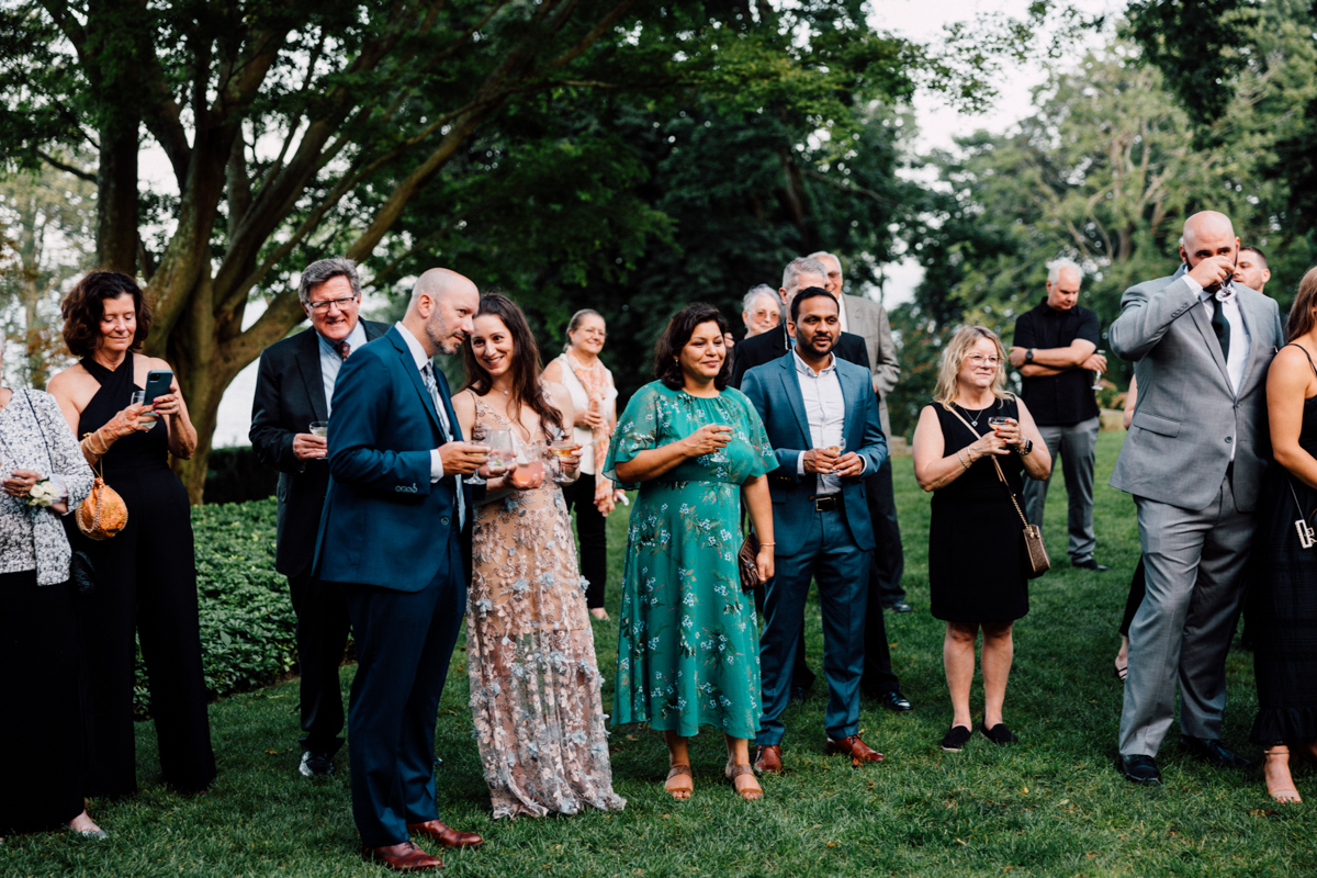 A diverse group of well-dressed individuals enjoying drinks outdoors at a social event, surrounded by greenery.