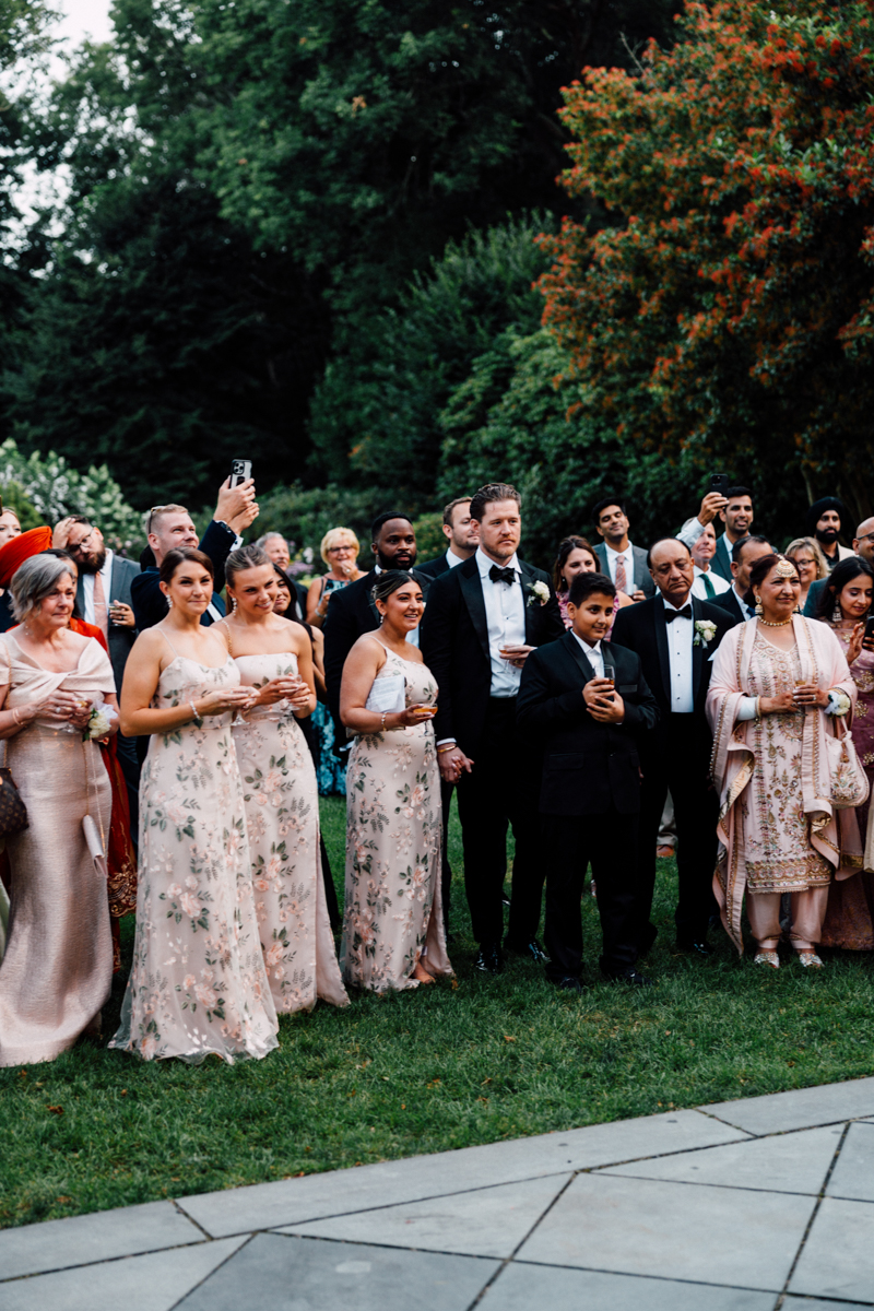A diverse group of wedding guests enjoying a beautiful outdoor celebration, dressed in formal attire, surrounded by lush greenery.
