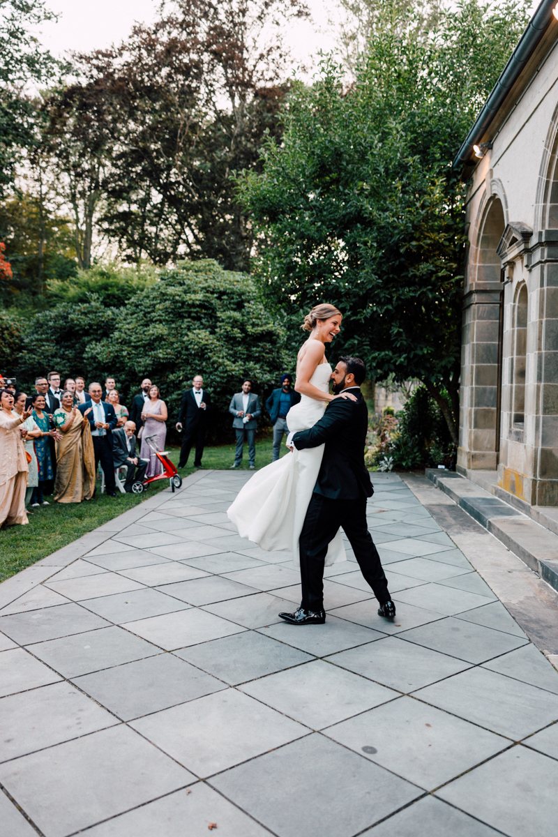 A groom lifts his bride in the air during an outdoor wedding celebration, surrounded by smiling guests in formal attire.