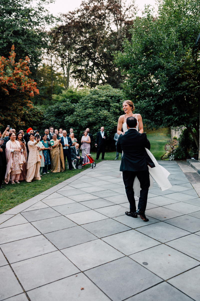 A groom lifts his bride as they share a joyful moment on the dance floor, surrounded by smiling guests in formal attire enjoying the celebration outdoors.