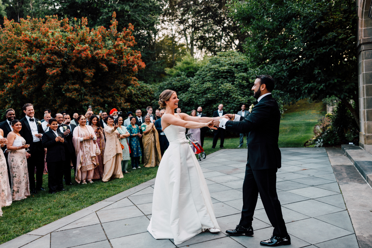 A joyful couple dances together at their wedding, surrounded by an enthusiastic group of guests in formal attire, set in a beautiful outdoor venue.
