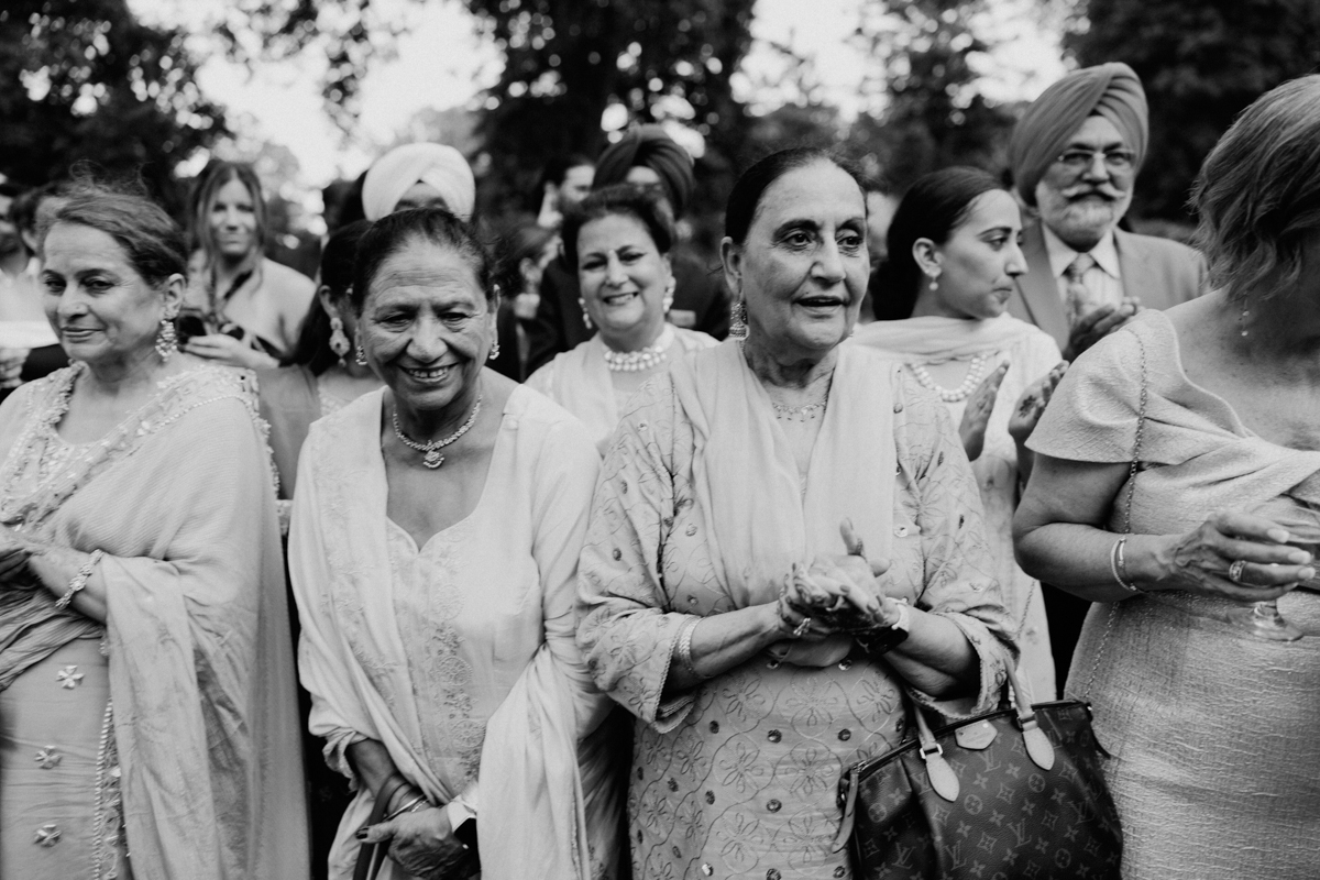 A group of women dressed in elegant attire smiling and enjoying a lively gathering, with a mix of traditional and contemporary styles in a festive outdoor setting.