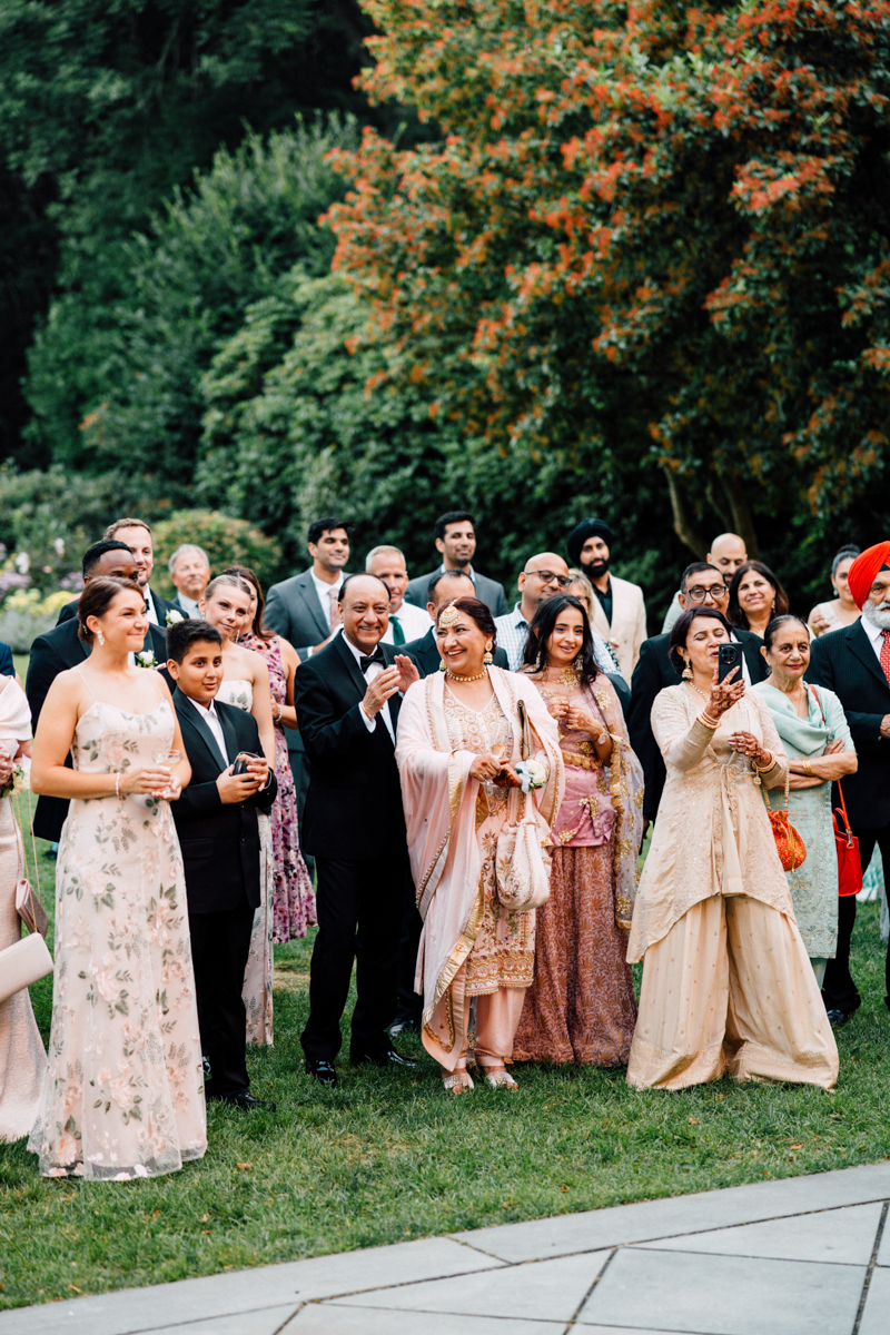 A diverse group of guests joyfully engaged in conversation while holding drinks at an outdoor event, surrounded by greenery and vibrant colors of autumn.