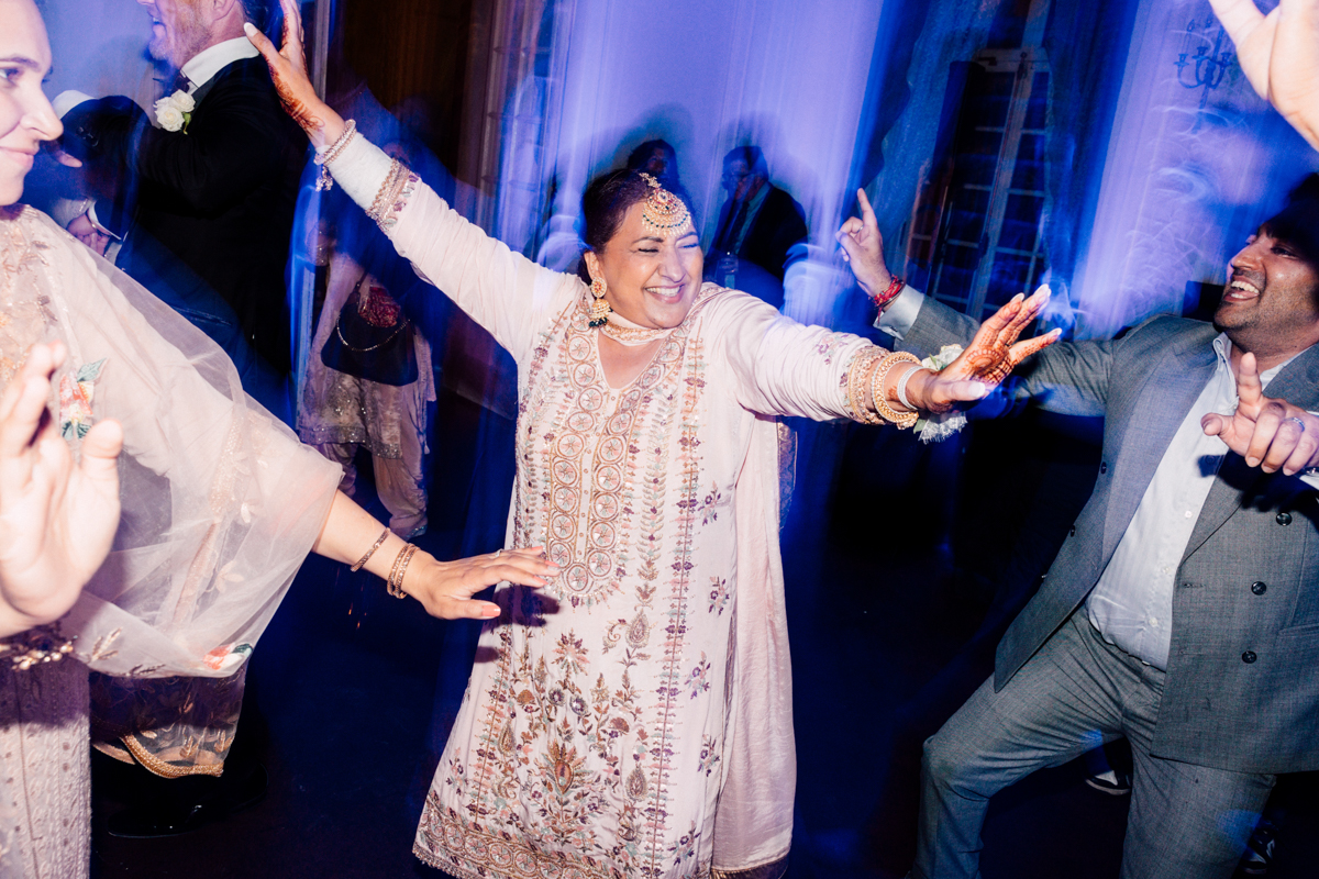 A woman in traditional attire joyfully dances at a lively event, surrounded by guests, in a vibrant, illuminated setting.