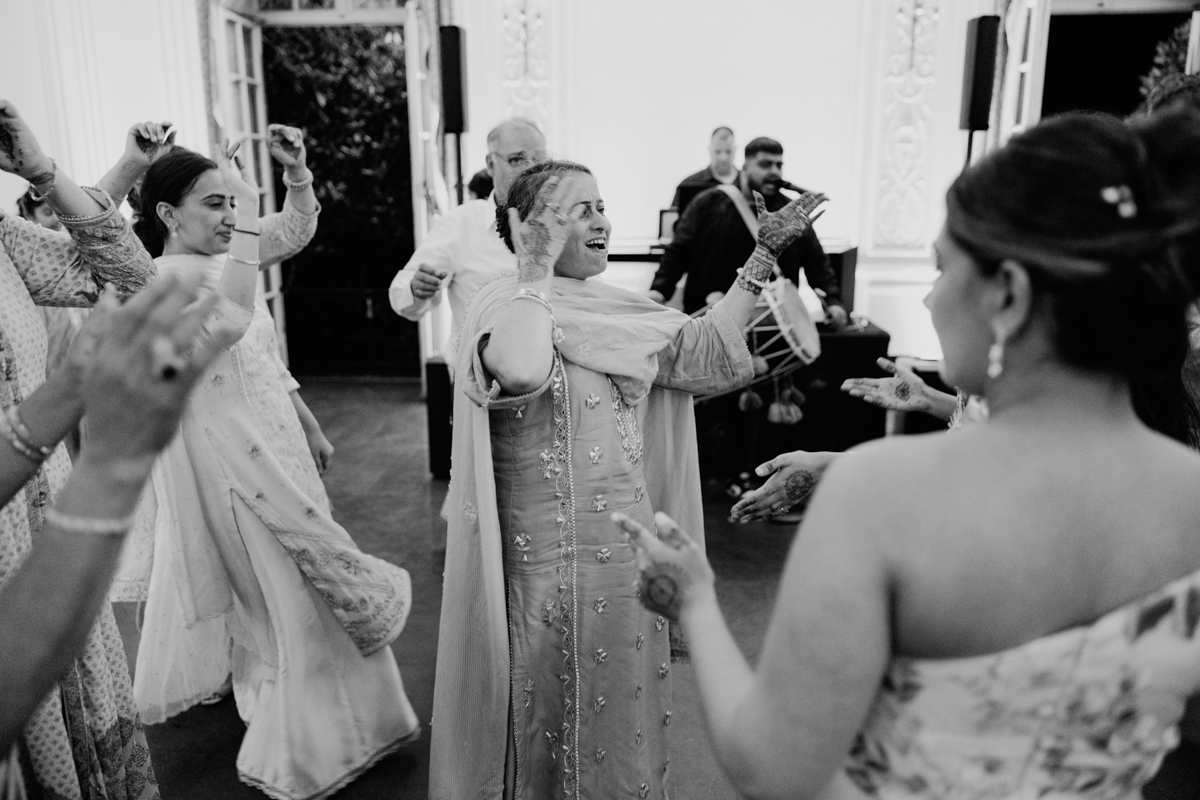 A group of people joyfully dancing together, adorned in traditional attire, with vibrant expressions and intricate henna designs, in a lively indoor setting.