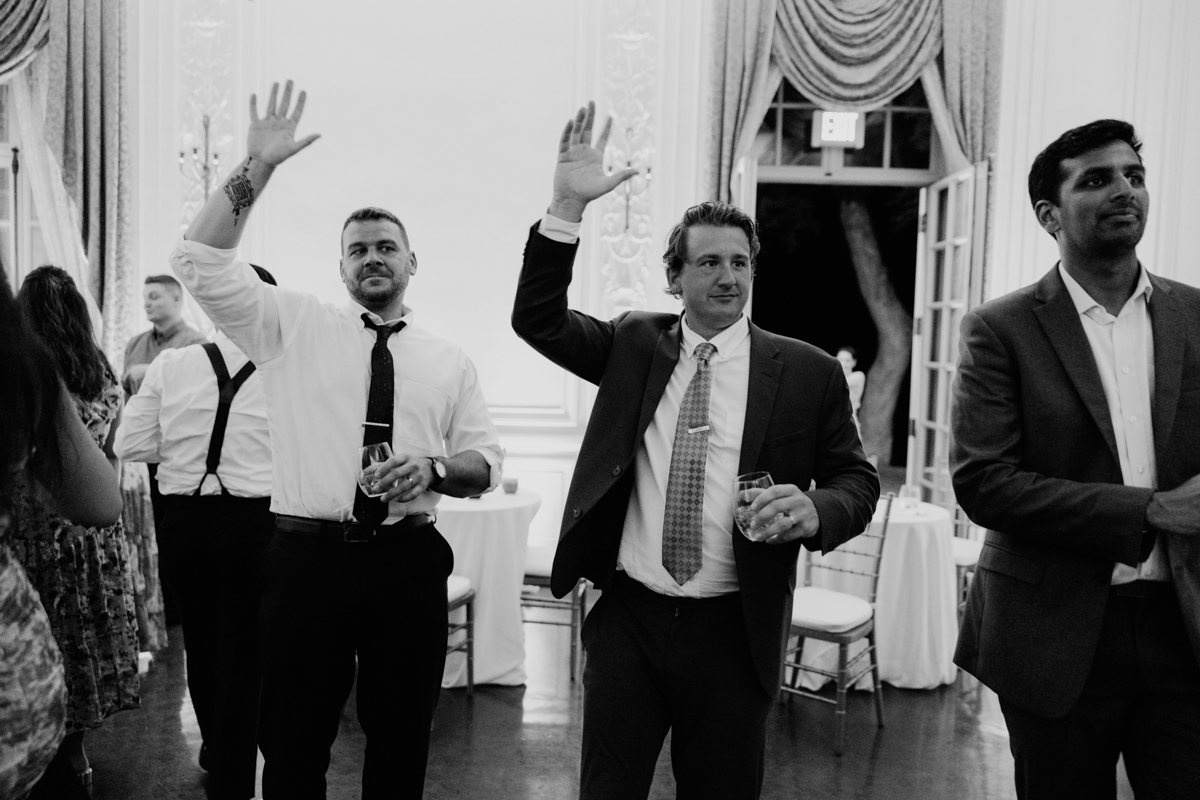 A group of well-dressed men at a lively event, raising their hands and holding drinks, with elegant decor in the background.