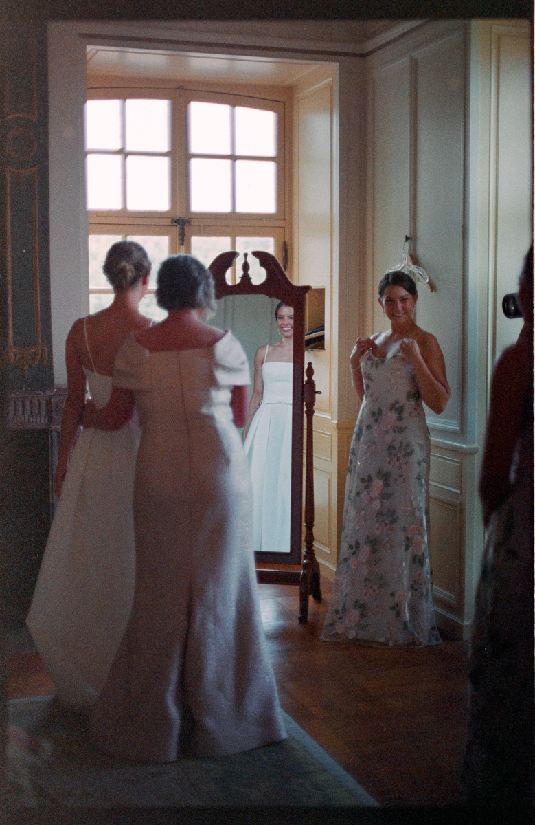 A bride admires her reflection in a vintage mirror while surrounded by friends, showcasing elegant wedding attire and a warm, intimate atmosphere.
