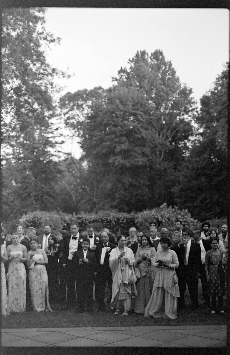 A large group of elegantly dressed individuals standing together in a garden setting, captured in black and white.
