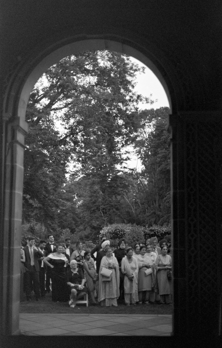A diverse group of people, dressed in formal attire, gather under an ornate archway, surrounded by lush greenery.
