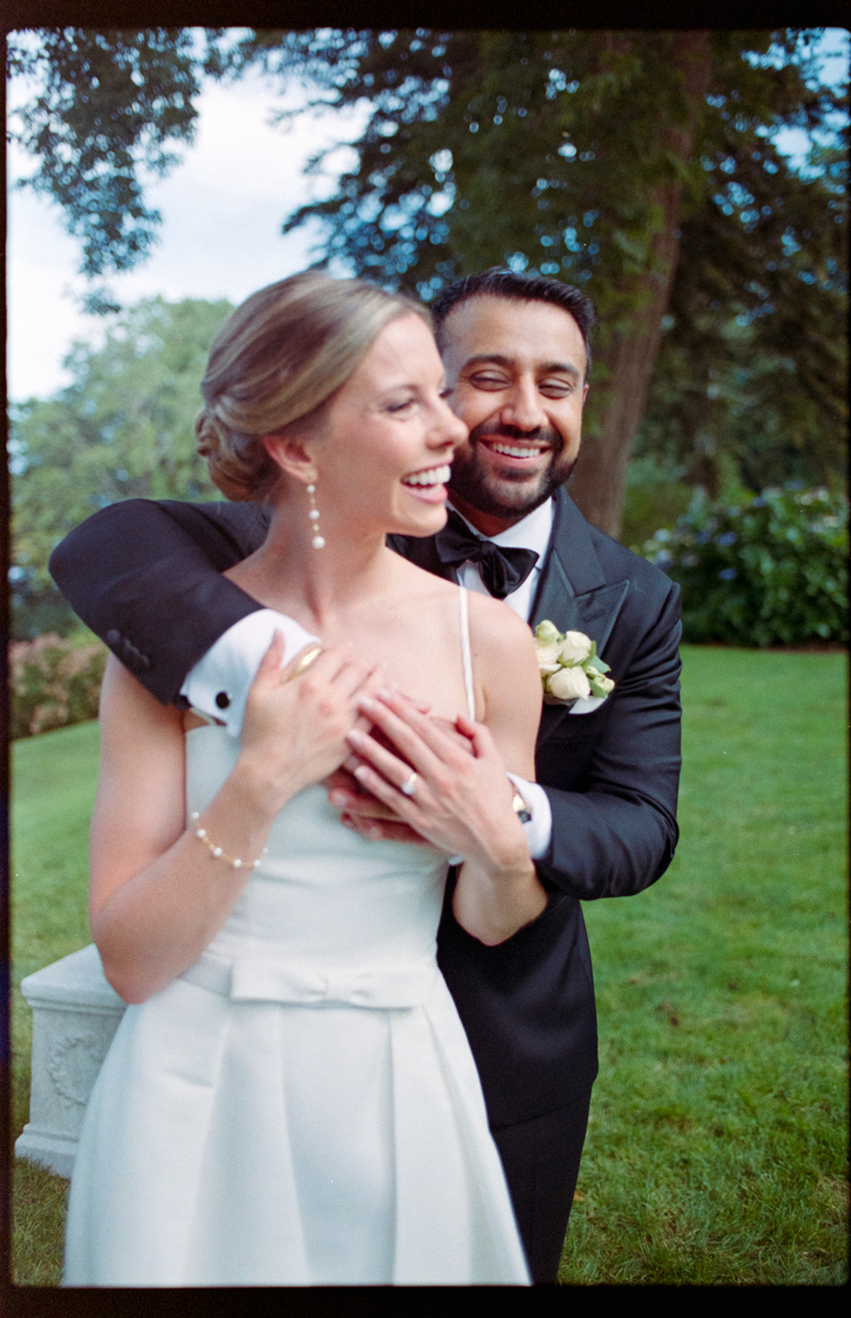 A smiling couple, dressed in wedding attire, share a loving embrace in a picturesque outdoor setting, surrounded by greenery.