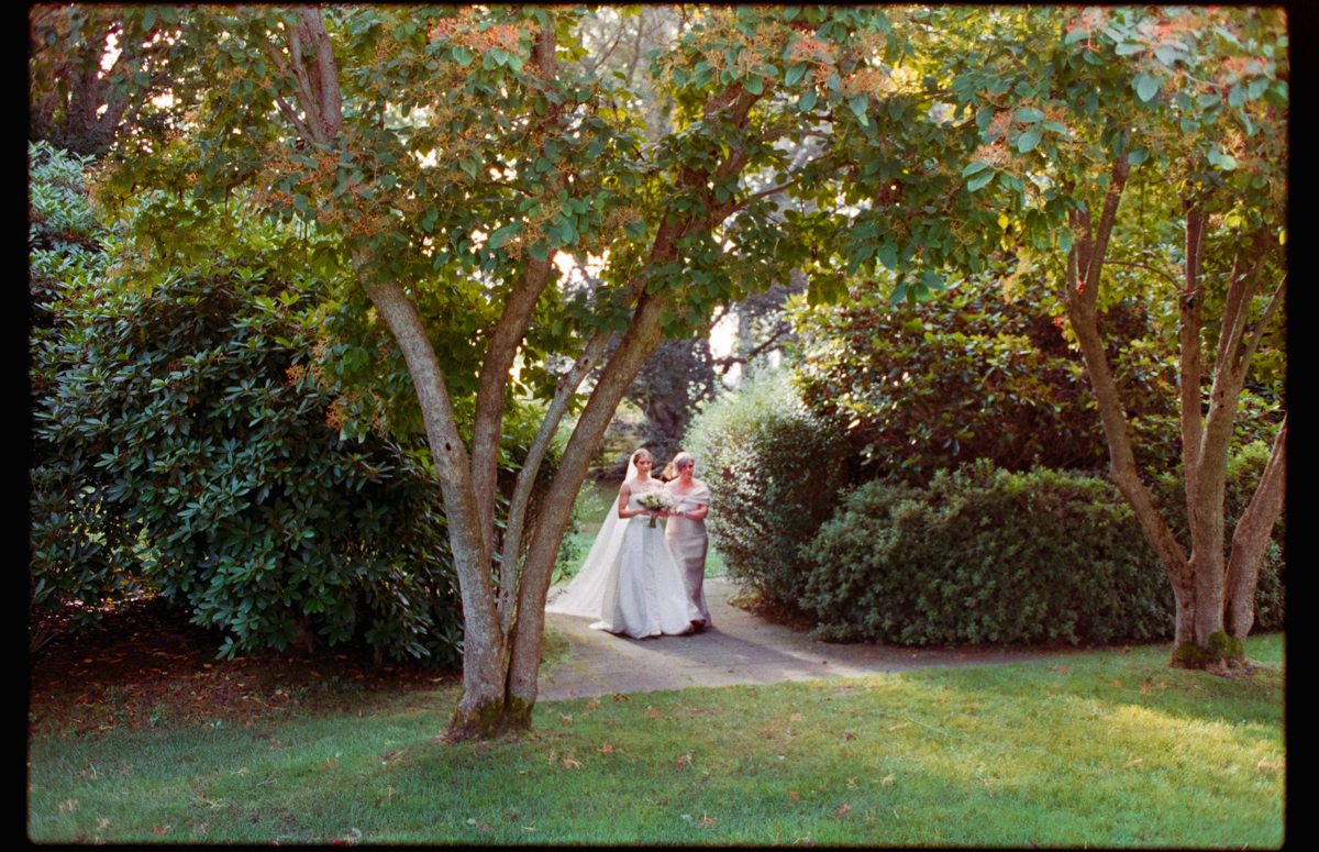 A bride and her mother walk together along a pathway surrounded by lush greenery, capturing a tender moment in a serene garden setting.