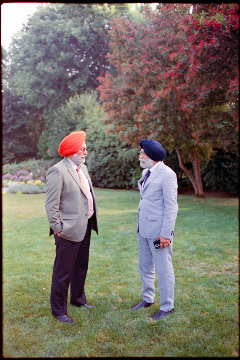 Two men wearing turbans, one orange and one blue, stand talking in a lush garden setting with greenery and colorful foliage in the background.