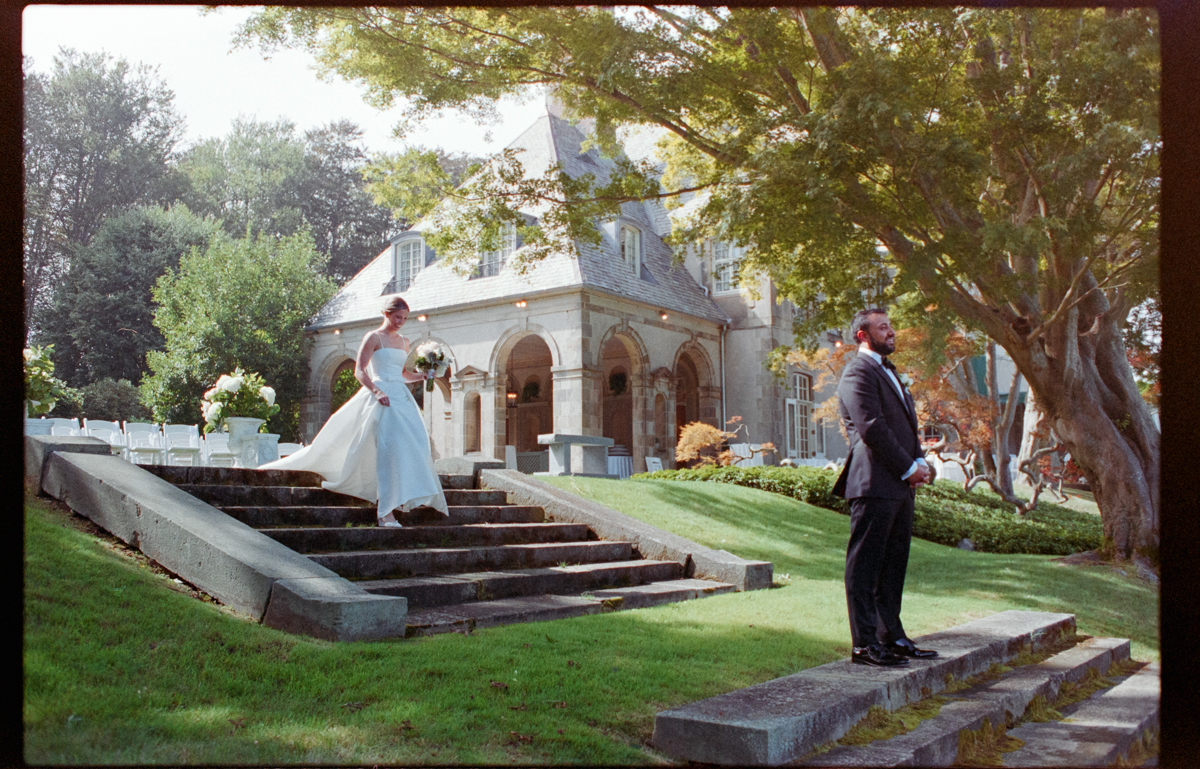 A bride descends stone steps towards a groom in a formal suit, set against an elegant garden venue with a historic mansion and lush greenery.
