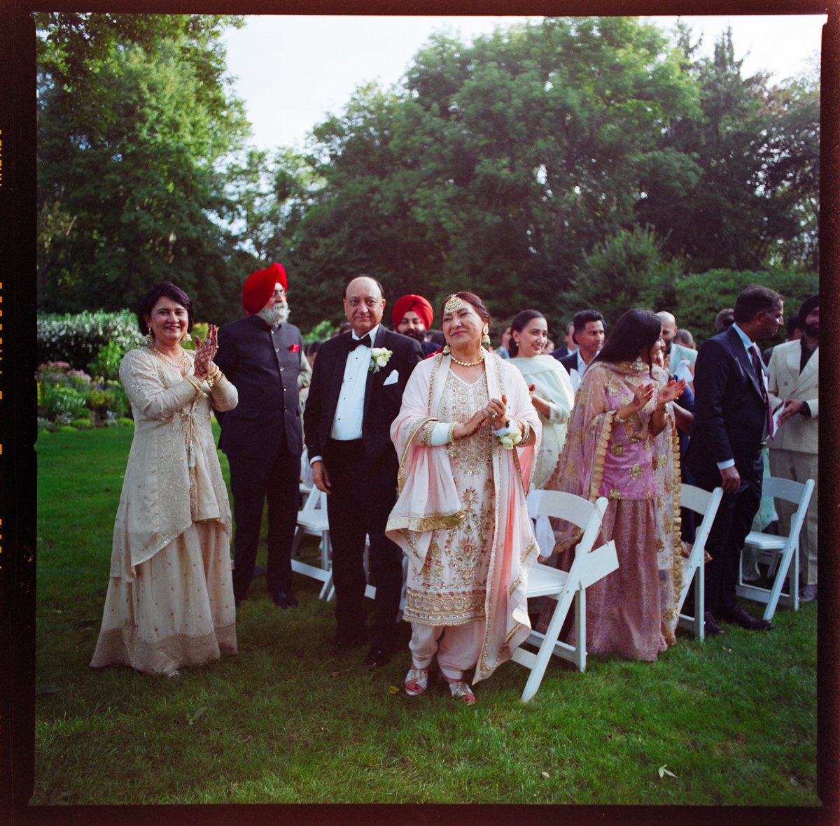 A joyful gathering of elegantly dressed guests clapping and smiling at an outdoor event, surrounded by lush greenery.