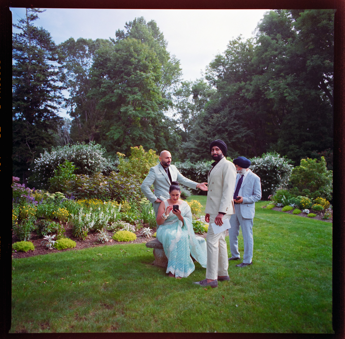Four people in formal attire gather on a garden lawn at Glen Manor House. One woman in a blue sari sits on a stone bench checking her phone, while three men in suits stand nearby, two focused on their phones amid blooming flowers and lush trees.